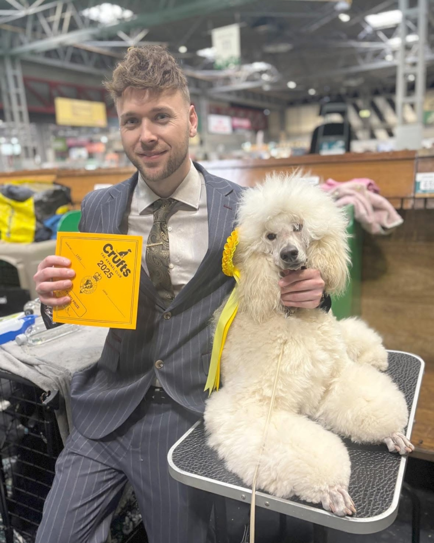 Our groomer in a suit at Crufts 2025 with Roisin, a groomed cream poodle, and a Crufts card